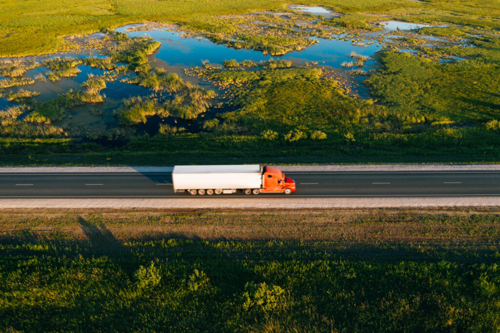 Semi truck with cargo trailer drive on road in green grass field with water at sunset, aerial drone view. Car with motion blur effect. Transportation background